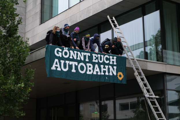 Menschen auf einem Gebäude mit einer Fahne, einem Baum links und Glasfenstern im Hintergrund, wahrscheinlich protestieren sie für die deutsche Regierung.
