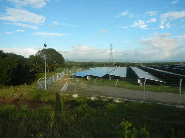 Ein großes Solarfeld in einem Feld, umgeben von einem Metallzaun, Bäumen, Pflanzen, Gras, Straßenmasten, Drähten und einem Turm.