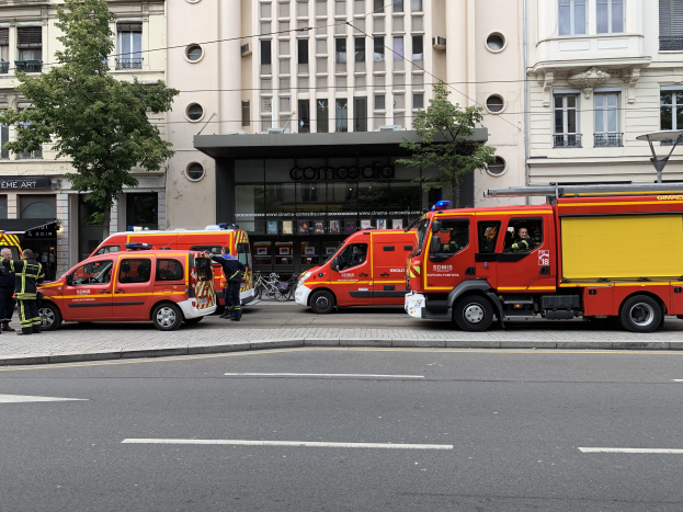 Eine Gruppe von Feuerwehrautos auf einer Straße in Paris geparkt, mit Menschen auf dem nahen Gehweg, Gebäuden, Bäumen und einem Fahrrad im Hintergrund.