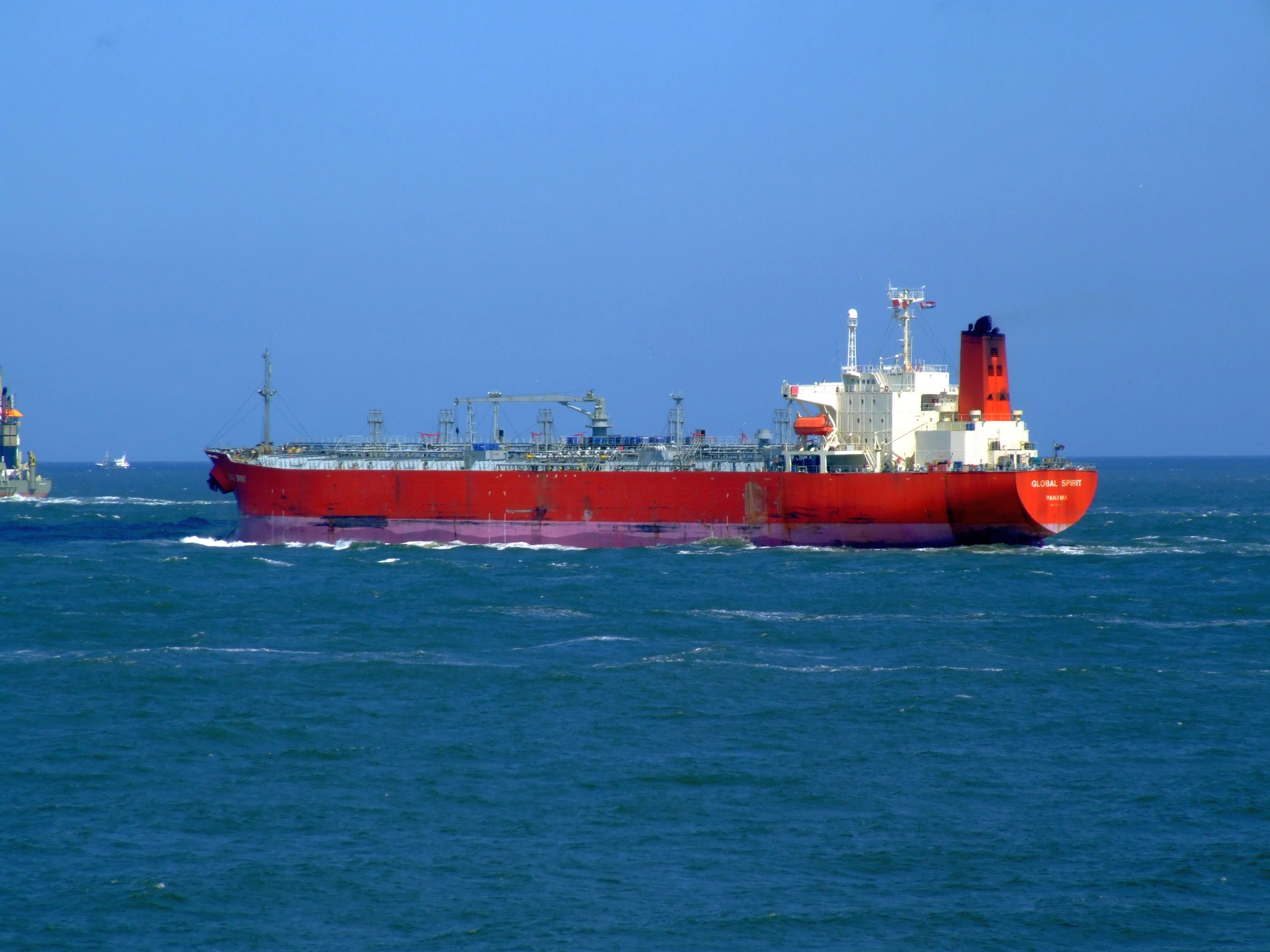 Großes rotes und weißes Öltankschiff in der Mitte des Ozeans mit anderen Schiffen im Hintergrund unter einem klaren blauen Himmel.