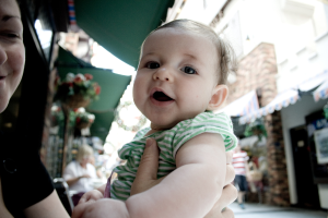 Eine Frau mit einem Kind auf dem Arm, im Hintergrund eine sitzende Frau, ein gehender Mann, Hauspflanzen, Gebäude und andere Gegenstände.