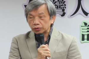 Ein Mann im Anzug spricht vor einer textbedeckten Wand in ein Mikrofon und sieht ernst aus.