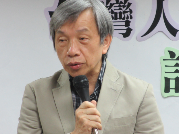 Ein Mann im Anzug spricht vor einer textbedeckten Wand in ein Mikrofon und sieht ernst aus.