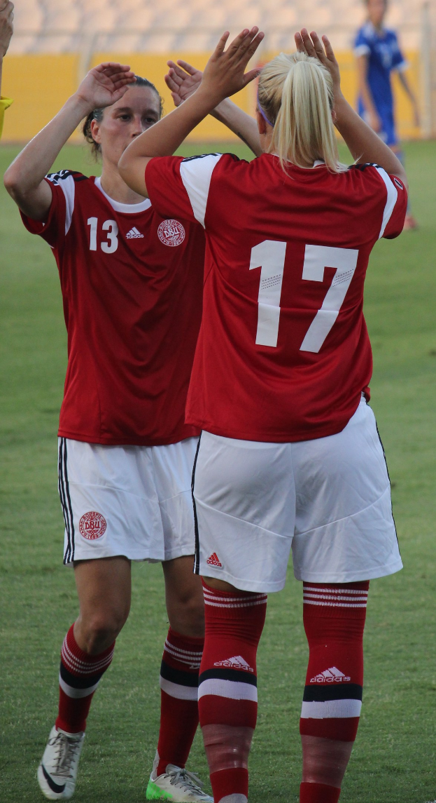 Zwei Frauen in roten Hemden und weißen Shorts klatschen auf einem Fußballfeld, mit einer Person in einem blauen Kleid, einer gelben Tafel und Stühlen im Hintergrund.