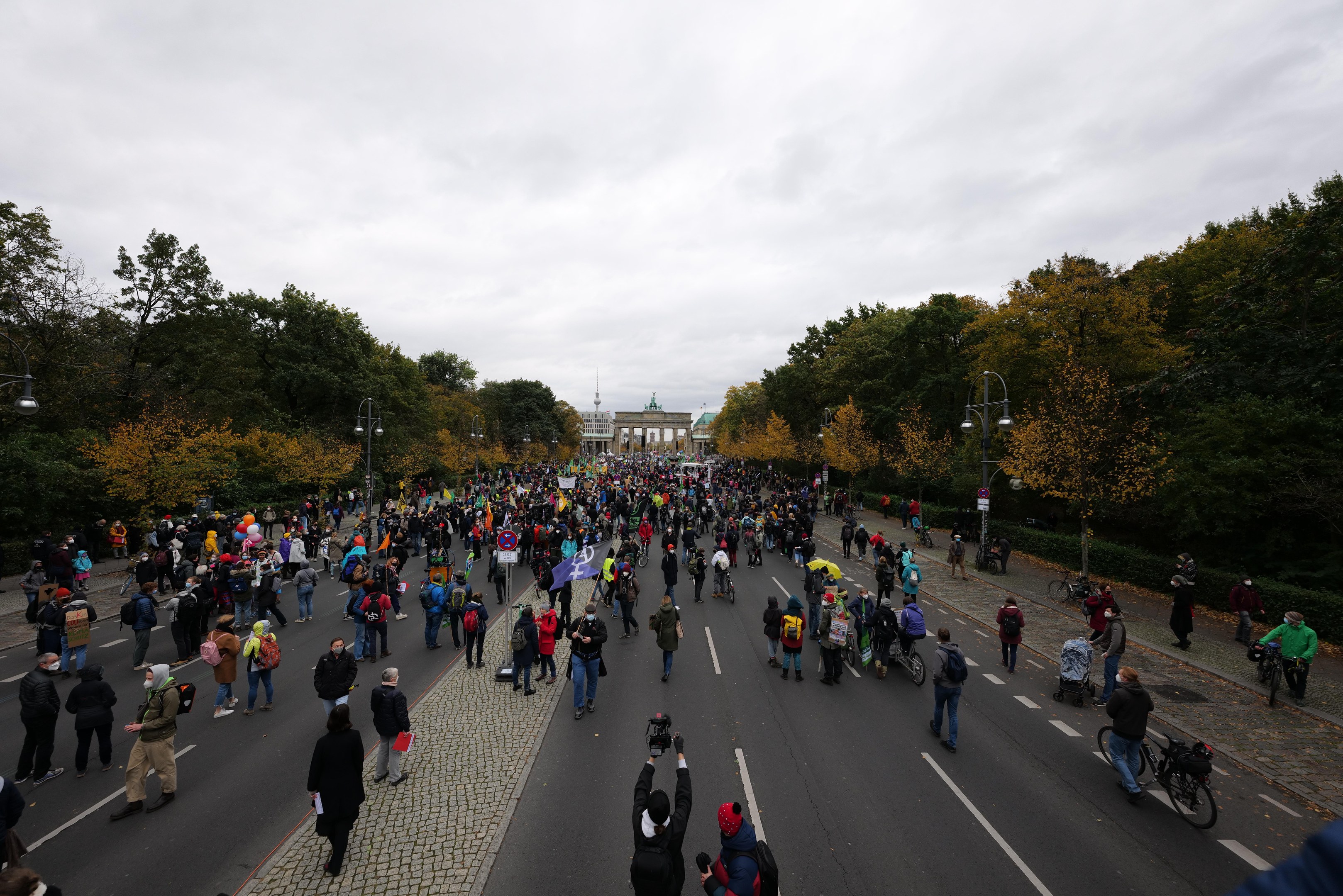 Eine große Gruppe von Menschen marschiert auf einer von Bäumen gesäumten Straße in Berlin, hält Kameras und hat ein Gebäude und einen klaren Himmel im Hintergrund.