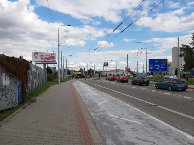 Eine Straßen in der Stadt mit Autos, einem Gehweg auf der linken Seite, einer Wand mit Graffiti, Laternen, Bäumen, Gebäuden und einem bewölkten Himmel.
