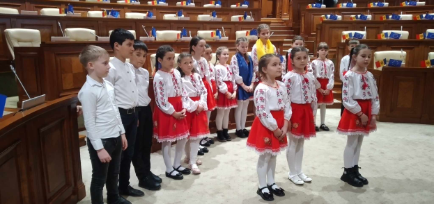 Eine Gruppe von Kindern in weißen und roten Kleidern steht zusammen in einem Raum, mit einem Podium und einem Mikrofon auf der linken Seite und Stühlen und Tischen mit verschiedenen Gegenständen im Hintergrund, die Fahnen tragen, die ihre Teilnahme an einer Nationalversammlung anzeigen.