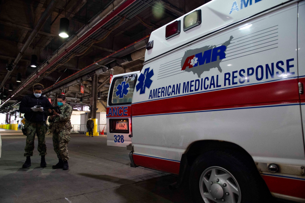 Ein Rettungswagen der American Medical Response (AMR) steht in einem Gebäude, vor ihm stehen zwei maskierte Personen; auf der linken Seite ist ein Fahrzeug zu sehen und im Hintergrund gibt es Menschen, Lichter, Rohre und andere Gegenstände.