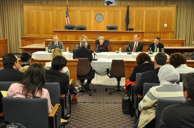 Ein Konferenzraum mit einer Gruppe von Menschen, die auf Stühlen sitzen, zwei ausländischen Flaggen, einem orangen Schreibtisch mit Wasserflaschen und einem Mikrofon.