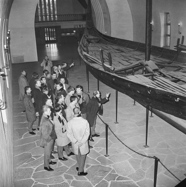Eine Gruppe von Menschen steht um ein Wikingerboot in einem Museum herum, das Boot ist rechts, Stangen und Seile davor und eine beleuchtete Wand links, alles in Schwarz-Wei├č.