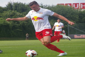 Ein Mann in einem Fußballtrikot kickt einen Ball auf einem grasbewachsenen Feld mit Bäumen und einem klaren blauen Himmel im Hintergrund, wobei ein "RB Leipzig"-Schild sichtbar ist.