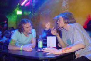 Ein Tisch mit einer Flasche, Speisekarte und Aschenbecher in der Mitte einer Indoor-Einrichtung, mit drei Personen, die Zigaretten rauchen.