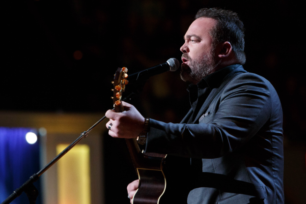 Ein Mann in einem Anzug spielt Gitarre und singt in ein Mikrofon auf der Bühne, mit Dierks Bentley im Hintergrund.
