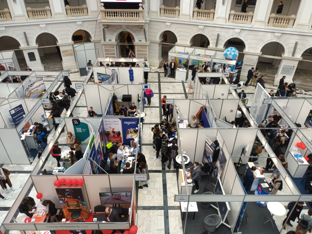 Eine große Gruppe von Menschen versammelt sich in einem Raum, der mit Ständen, Bannern, Tischen, Stühlen und Luftballons gefüllt ist, mit einem Gebäude im Hintergrund, das Bögen, Säulen, Geländer und einen Bildschirm aufweist.
