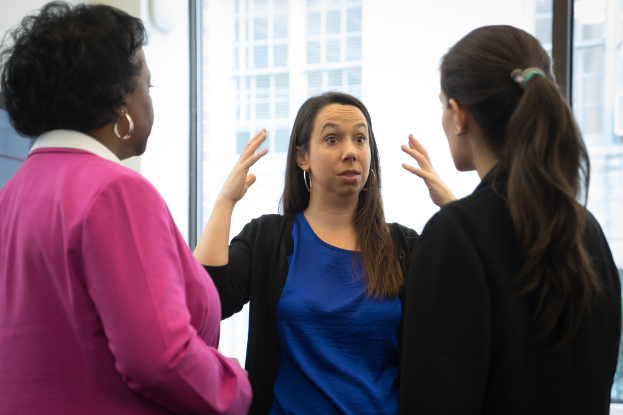 Drei Frauen, die stehen, eine in einer schwarzen Jacke, die erklärt, mit durchsichtigen Gläsern und einem Gebäude im Hintergrund.