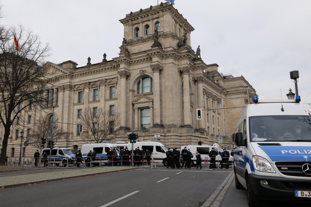 Eine Gruppe von Polizisten steht vor dem Reichstagsgebäude in Berlin, Deutschland, mit Fahrzeugen, einem Zaun, Verkehrszeichen, Laternen, Bäumen und Flaggen im Hintergrund, bei klarem Himmel.