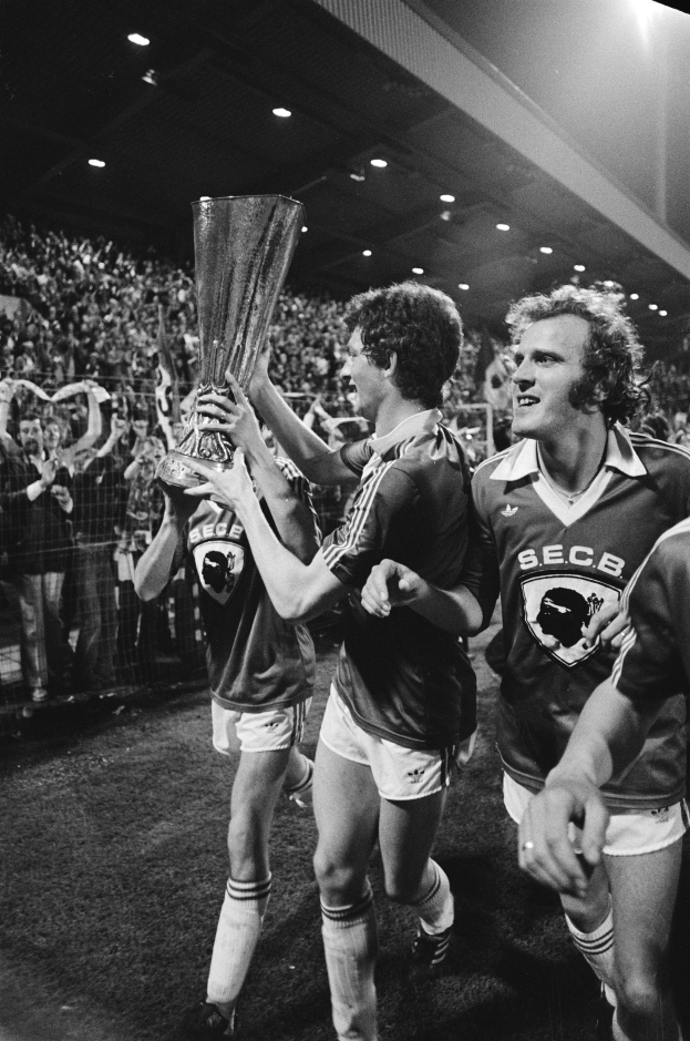 Schwarzes und weißes Bild von Fußballspielern, die einen Pokal auf einem Feld feiern, umgeben von einer Menge hinter einem Zaun unter Stadionbeleuchtung.