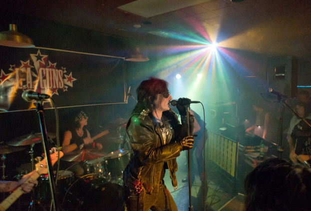 Eine Frau in einer schwarzen Jacke singt in ein Mikrofon auf der Bühne, mit einem Mann in Sonnenbrille und Stirnbinde, der ein Instrument spielt, und einem Plakat mit "LA Guns" im Hintergrund, unter Diskokugeln und Hängelampen.
