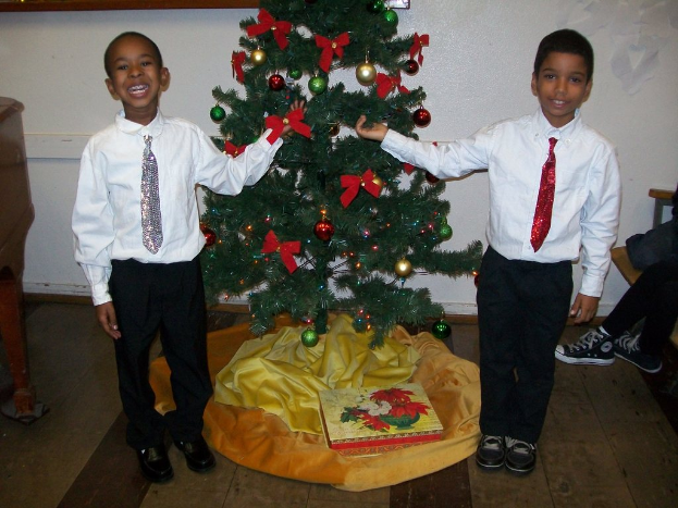 Ein geschmückter Weihnachtsbaum steht in einem Raum mit einem Holztisch links, einem Stuhl rechts und zwei Jungen in weißen Hemden, die strahlen und den Baum halten.
