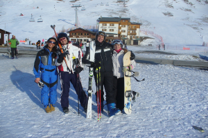 Vier Personen posieren für ein Foto mit Skate-Pads und Werkzeugen, mit Gebäuden und anderen Menschen im Hintergrund, vor einer schneebedeckten und vereisten Berglandschaft.