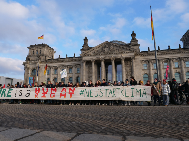 Eine Gruppe von Menschen steht vor dem Reichstagsgebäude in Berlin, Deutschland, mit einer Tafel, auf der "Wir sind ein Menschenrecht" steht, während das Gebäude mit Säulen, Fenstern, Bögen und Statuen geschmückt ist und von Fahnen umgeben ist.