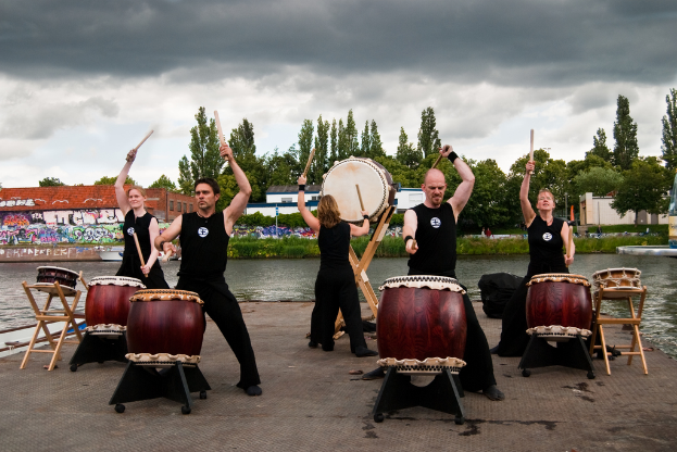 Fünf Menschen in schwarzen Kleidern spielen Trommeln auf einem kleinen Stuhl an einem offenen Bereich umgeben von Wasser, Bäumen und einem bewölkten Himmel, mit einem Gebäude im Hintergrund.
