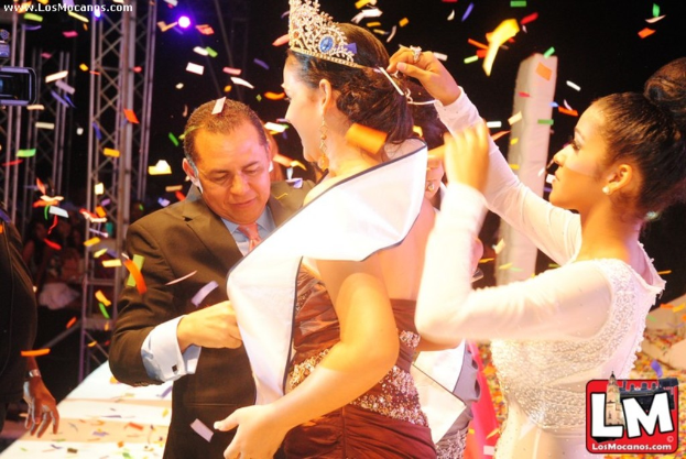 Ein Mädchen auf der Bühne mit einer Krone, während ein anderer Mädchen sie anpasst und ein Mann im schwarzen Anzug ihr die Hand schüttelt, mit Metallrahmen und dunklem Hintergrund.