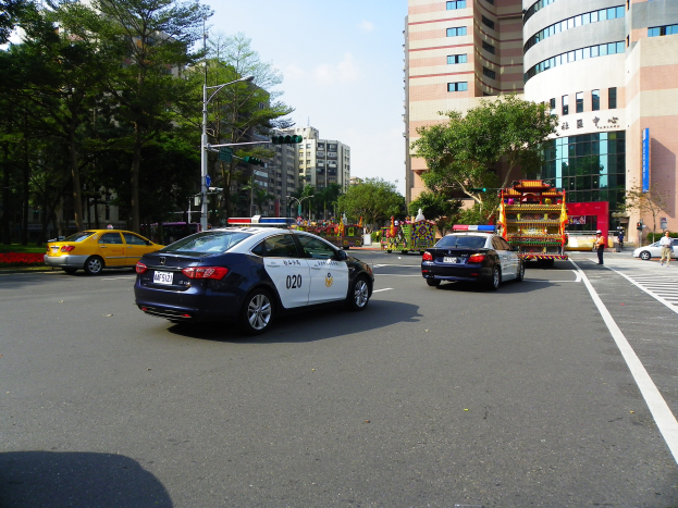 Polizeiauto fährt auf einer städtischen Straße mit hohen Gebäuden, Bäumen, Straßenlaternen, Ampeln und Fußgängern unter einem bewölkten Himmel.