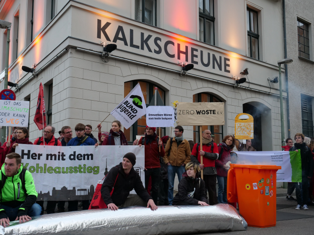 Eine Gruppe von Menschen hält Schilder und Plakate vor einem Gebäude während einer Demonstration in Deutschland, mit zwei Personen auf einem Objekt sitzend im Vordergrund und einem Müllcontainer auf der rechten Seite.