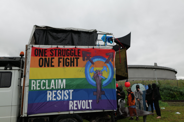 Eine Gruppe von Menschen steht vor einem Lkw mit einer Fahne, auf der "Ein Kampf, ein Kampf, zurückerobern, widerstehen, revoltieren" steht, der auf einem grasbewachsenen Gebiet mit Pflanzen und einem Gebäude im Hintergrund unter einem klaren blauen Himmel geparkt ist.