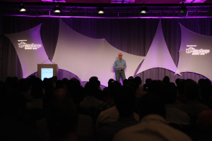 Ein Mann in einem blauen Hemd steht auf einer Bühne mit einem schwarzen und weißen Vorhang dahinter und spricht zu einem Publikum, das vor ihm sitzt.