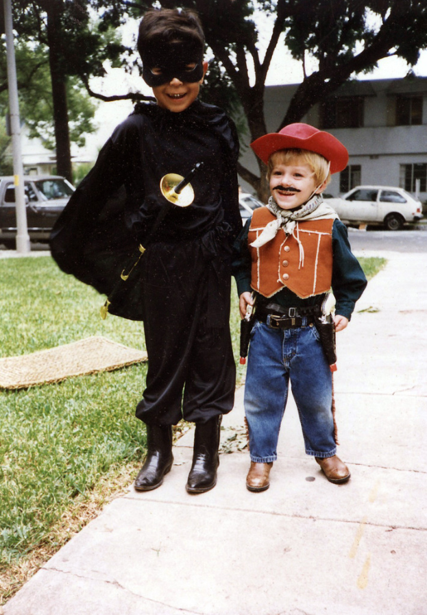 Kinder in verschiedenen Kost√ľmen stehen auf einer Stra√üe mit Geb√§uden, B√§umen und Fahrzeugen im Hintergrund.