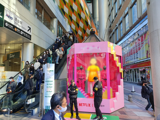 Eine Gruppe von Menschen steht vor einem Gebäude mit Glaswänden, Säulen und Texttafeln, einige tragen Masken, während eine Rolltreppe mit Menschen daneben fährt; eine pinkfarbene Box in der Mitte zeigt Schaufensterpuppen und trägt die Aufschrift "Netflix's new Netflix booth in Tokyo."
