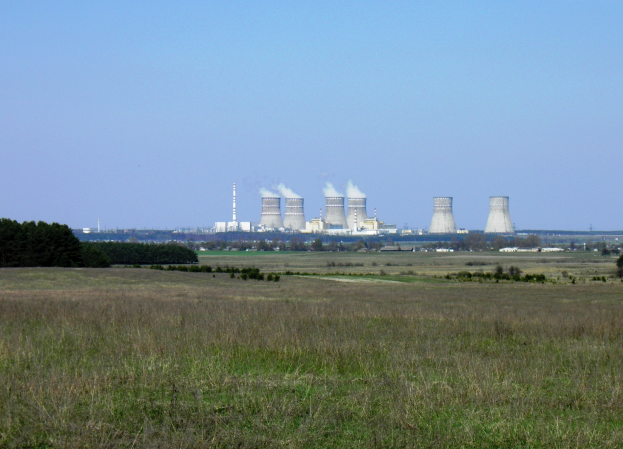 Ein Atomkraftwerk in einer von Bäumen umgebenen Wiese, aus dessen Schornsteinen Rauch aufsteigt und der Himmel im Hintergrund zu sehen ist.