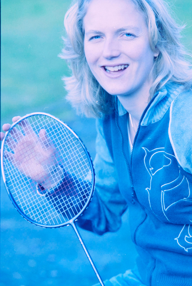 Eine Frau mit strahlendem Lachen, die ein weißes Hemd und blaue Shorts trägt, hält einen Badminton-Schläger und schaut konzentriert darauf, bereit zum Spiel.
