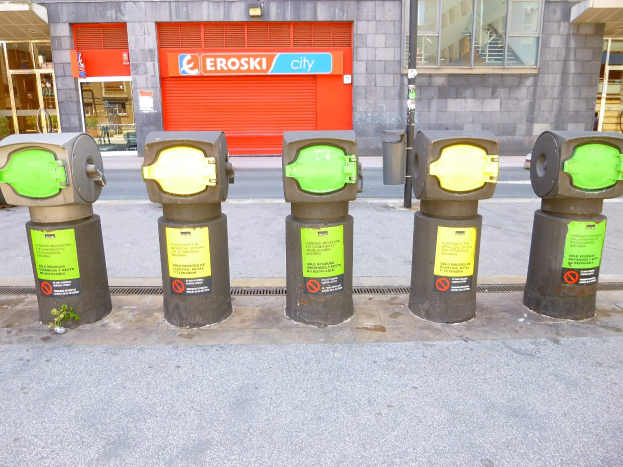 Eine Reihe von Parkautomaten an einer Stadtstraße, mit einem Gebäude mit Glasfenstern und einer Rolllade mit Text im Hintergrund und einem nahen Müllcontainer.