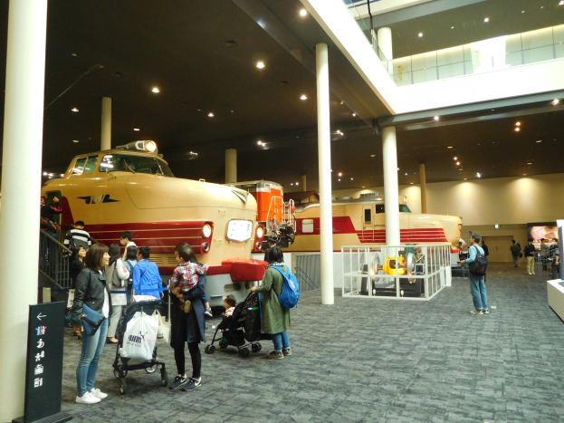 Menschen vor einem Zugmodell in einem Museum mit Säulen, Geländern und Deckenleuchten, einige mit Taschen oder Kinderwagen und andere die Ausstellungen beobachtend, mit einem Textschild links sichtbar.