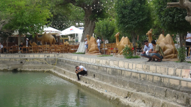 Ein Mann sitzt am Rand eines Teiches umgeben von Menschen, Bäumen, Skulpturen, Möbeln und Schirmen, mit Wasser im Vordergrund und Himmel im Hintergrund.