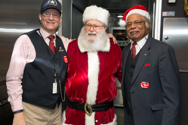 Eine Gruppe von Menschen mit Brillen, Mützen, Krawatten, ID-Karten und Abzeichen, wobei einer ein Weihnachtskostüm trägt, vor einer Stahlwand mit Lichtern, Schildern und anderen Gegenständen im Hintergrund.
