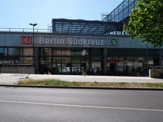 Große Glaswand-Flughafen Berlin Südkreuz mit sichtbarem Text, umgeben von Straßeninfrastruktur, Fahrzeugen, Fütter, Fahrrädern, Bäumen und einem klaren Himmel.