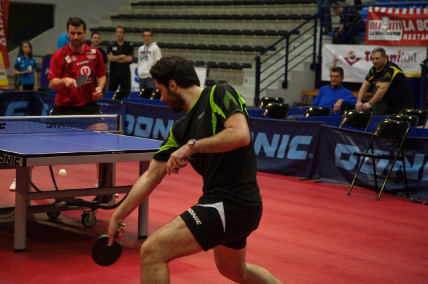 Eine Gruppe von Menschen in verschiedenen Farben, darunter schwarz, weiß und blau, sitzt um einen Tisch mit einem Spiel, Stühlen und einer Treppe im Hintergrund.