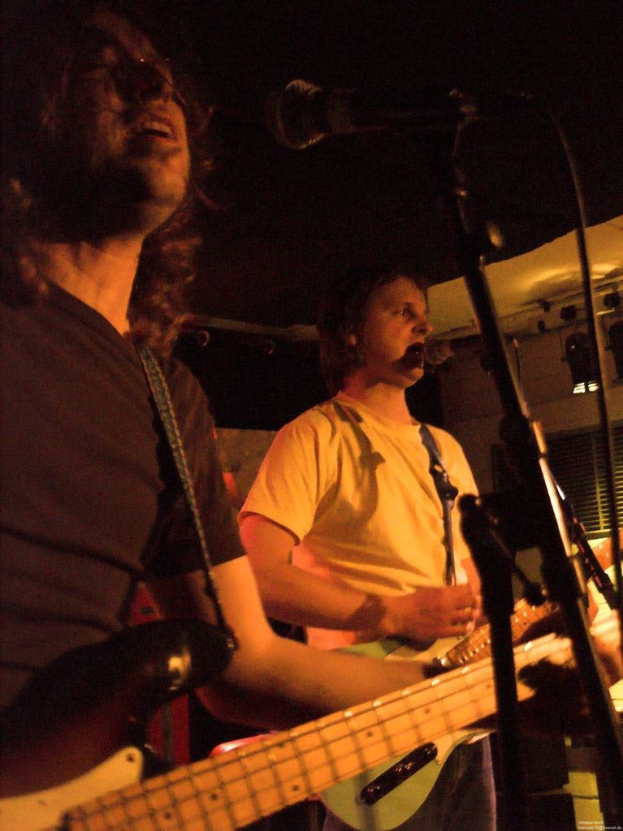 Eine Person in einem wei├čen Hemd singt in ein Mikrofon und spielt eine grüne Gitarre, steht neben einer anderen Person in einem schwarzen Hemd, Brille und spielt eine schwarze Gitarre, mit Scheinwerfern und Musikinstrumenten im Hintergrund.