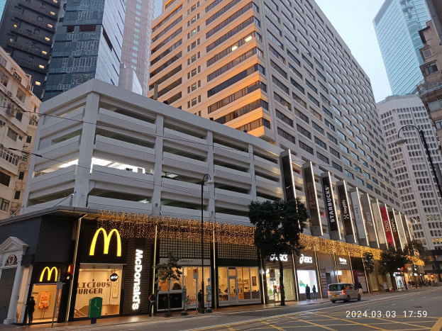 Ein McDonald's-Restaurant auf einer belebten Stadtstraße, umgeben von hohen Gebäuden, Fahrzeugen, Fußgängern, Straßenlaternen und Bäumen unter einem klaren blauen Himmel.