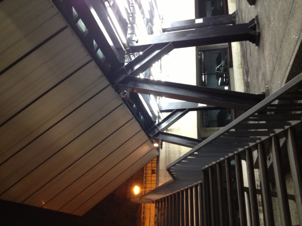 Treppen mit Geländern in der unteren rechten Ecke, Säulen auf der rechten Seite des Bodens, Autos in Gebäuden hinter den Säulen und Lampen auf der linken Seite mit sichtbarem Dach.