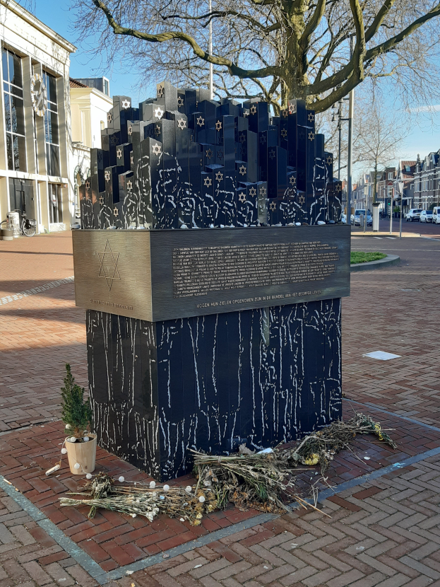 Ein Denkmal für Holocaust-Opfer steht auf einem Stadtplatz mit einem Baum davor, einem Topf mit einer Pflanze, verstreuten trockenen Blättern, Gebäuden, Fahrzeugen, Pfählen und Bäumen im Hintergrund.