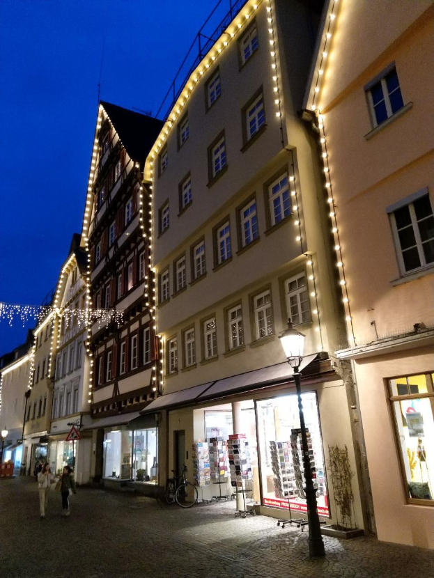 Headsteinstraße in Rothenburg ob der Tauber, Deutschland, geschmückt mit Weihnachtslichtern, mit Menschen, die spazieren gehen und ein Fahrrad in der Nähe geparkt, flankiert von Gebäuden mit Fenstern unter einem sichtbaren Himmel.