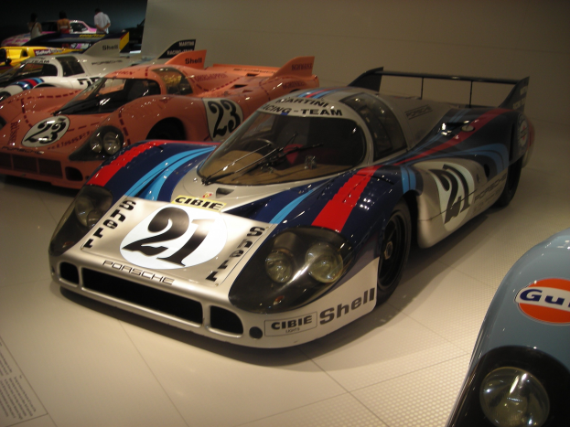 Ein Porsche 917K Rennwagen auf einer Museumsausstellung umgeben von anderen Sportwagen, mit Menschen in der Nähe und einer Wand im Hintergrund; ein Textschild ist auf der linken Seite zu sehen.