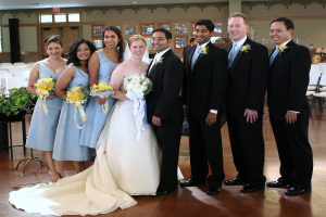 Eine Frau in einem weißen Kleid mit einem Strauß steht in der Mitte, umgeben von Frauen in blauen Kleidern mit Strauß und Männern in schwarzen Anzügen, die alle lächeln und drinnen mit verschiedenen Gegenständen im Hintergrund stehen.