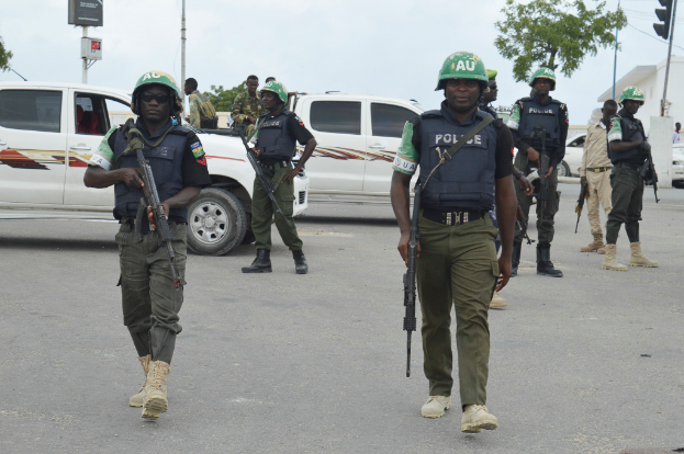 Eine Gruppe von Polizisten in Helmen, die Waffen halten, geht eine Straße entlang mit Fahrzeugen, Laternenmasten, Ampeln, Texttafeln, Bäumen, Gebäuden und einem bewölkten Himmel im Hintergrund.