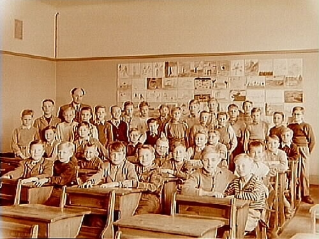 Ein altes Foto einer Klasse mit Kindern auf Bänken sitzend und auf dem Boden stehend, mit Papieren an der Rückwand geklebt.